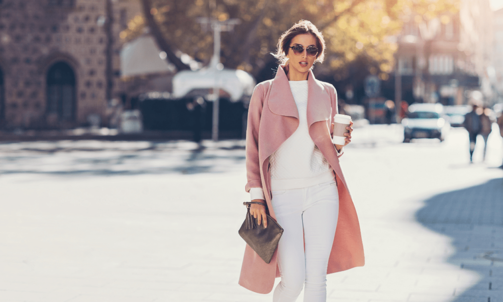 A fashionable woman wearing a pink trench coat, white outfit, sunglasses, and carrying a brown purse, holding a Starbucks coffee while walking down the street
