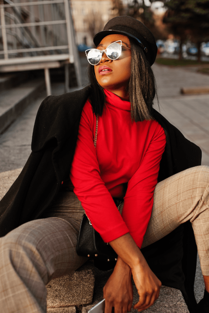 Woman stylishly seated, wearing sunglasses, a black blazer, and a vibrant red turtleneck, showcasing her versatile fashion choices.