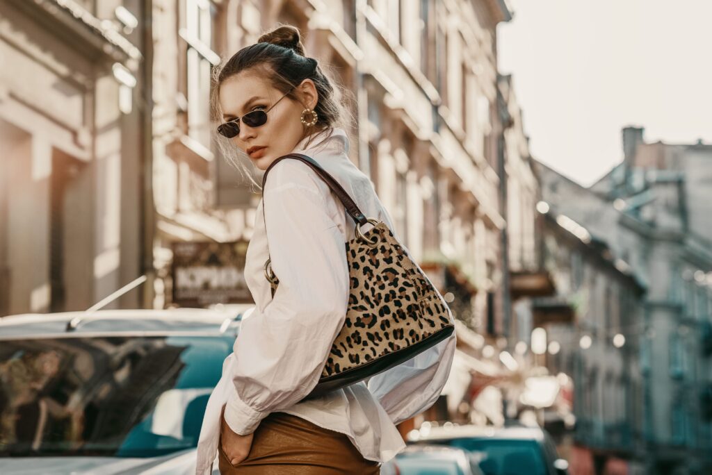 An outdoor fall fashion portrait of a stylish woman wearing sunglasses, a white shirt, leather trousers, and holding a leopard print bag, strolling through a European city with ample copy space