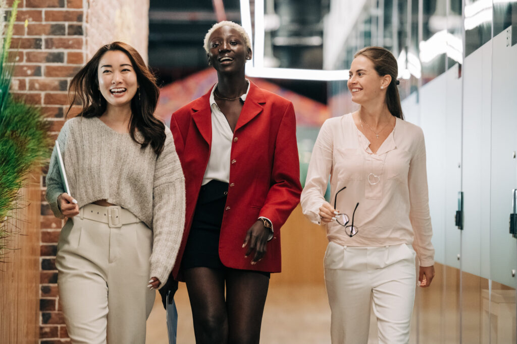 Three cheerful women strolling down the hallway in business casual attire, sharing smiles and enjoying each other's company.