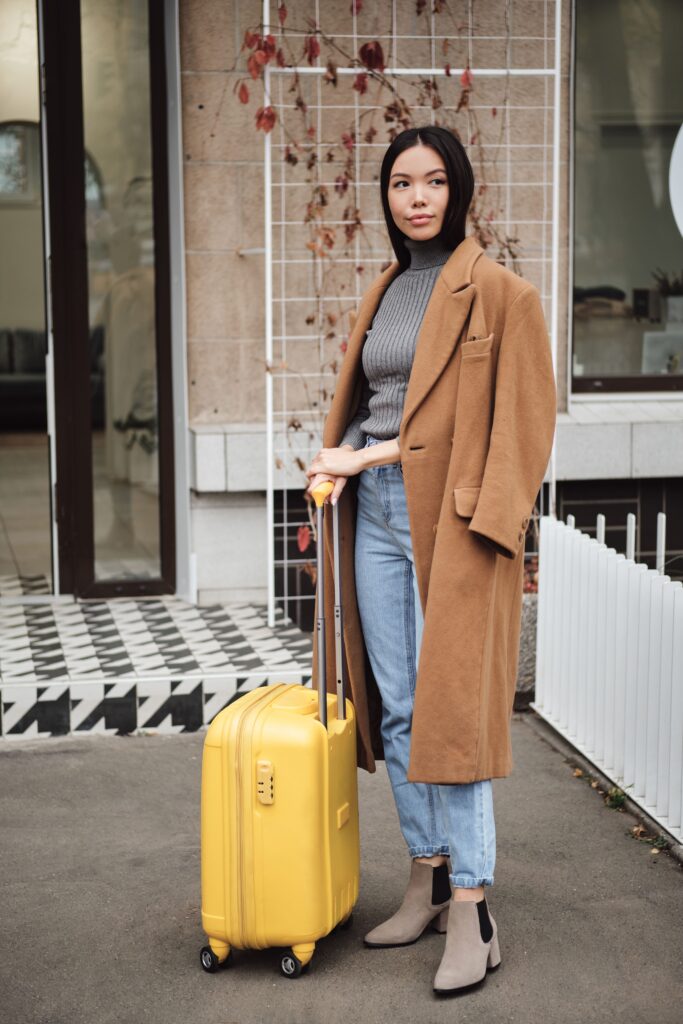Stylish Asian girl in a coat gazing dreamily while standing on a city street with a yellow suitcase.
