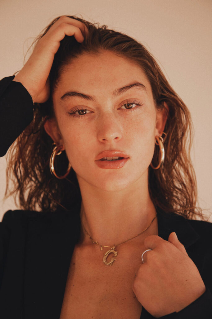 Freckled girl wearing earrings and a necklace, adorned in a black blazer with hands in her hair, strikes a self-portrait pose for a captivating facial close-up.