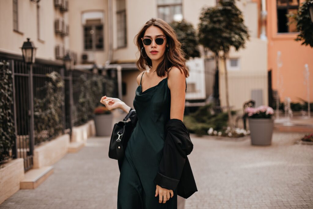 Fashionable pale brunette showcasing a capsule wardrobe outfit, wearing a long green dress, black jacket, and sunglasses, elegantly standing on a city street during daytime against a light-toned building wall.
