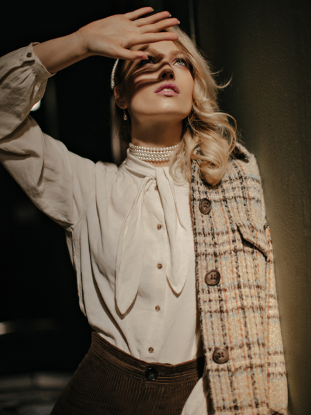 A blonde woman with curly hair wearing a tweed jacket and white blouse, posing in a dark room while covering the sun with her hand.