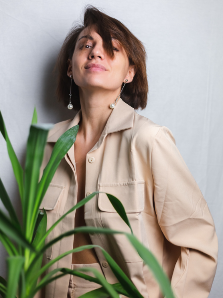 A serene indoor portrait featuring a gentle caucasian woman wearing a beige suit without a bra, posing behind a tropical palm plant, against a gray background.
