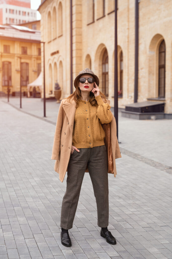 A self-assured young woman wearing an autumnal beige coat, sunglasses, and a plaid Panama hat stands tall in full height.