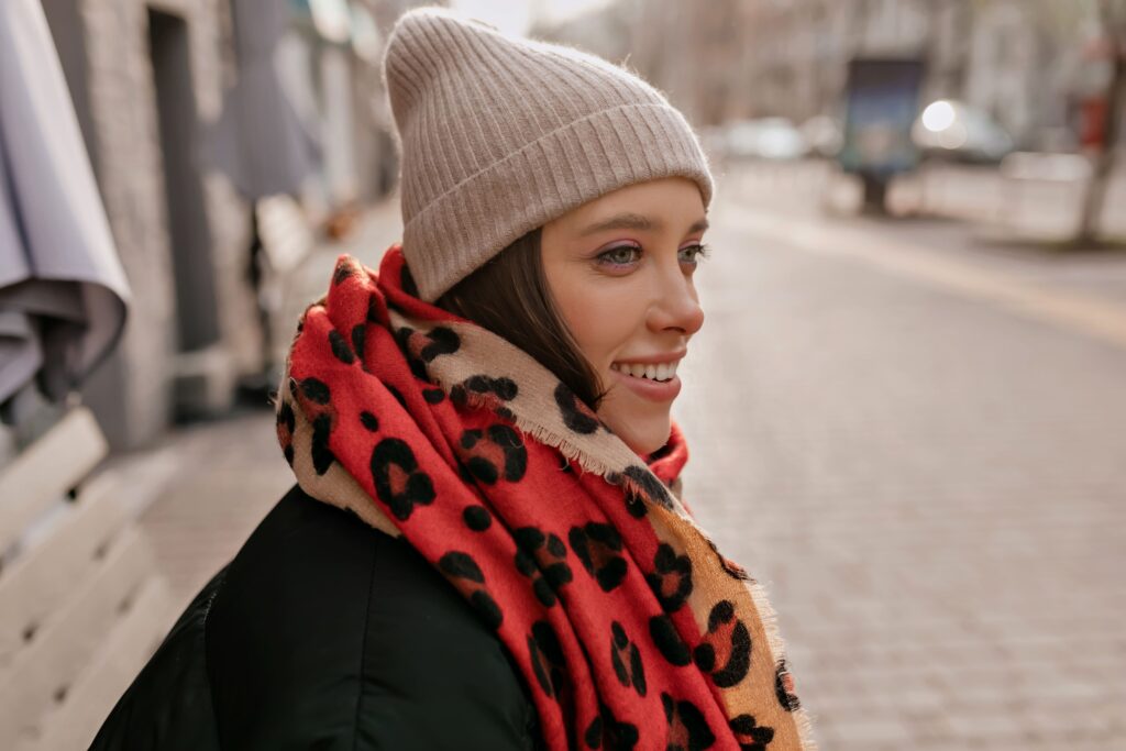 Complimentary image of a fashionable, grinning woman wearing a knit hat and vibrant scarf, seated on a bench outdoors in the city as she awaits her friends.