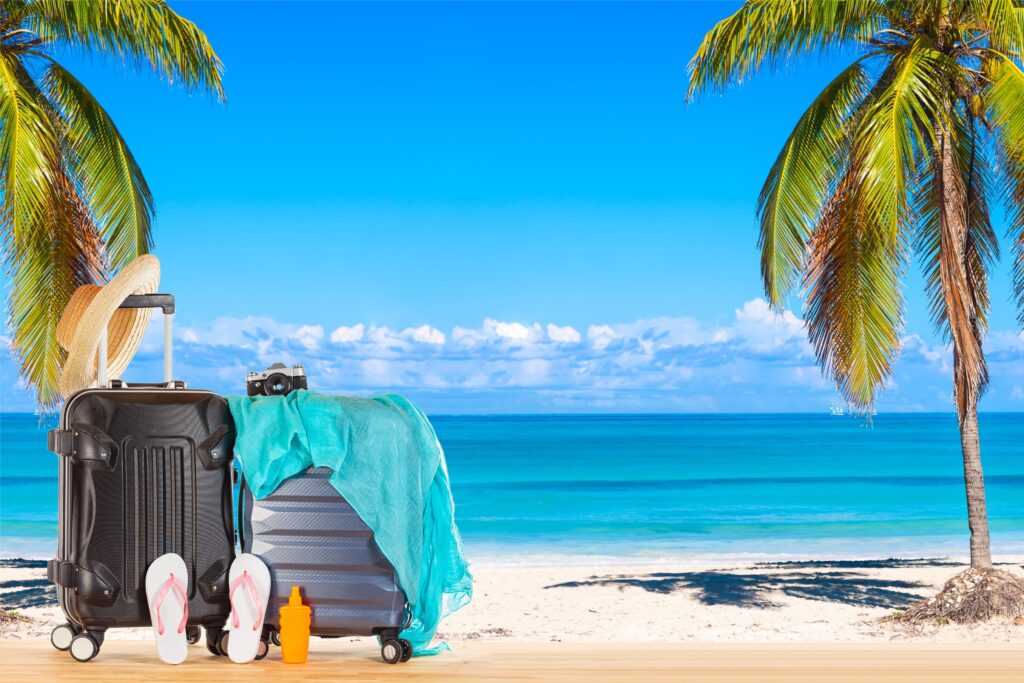 Photo suitcases baggage with straw hat, blue pareo, flip flops, sunscreen lotion bottle and retro camera against amazing ocean beach with palms