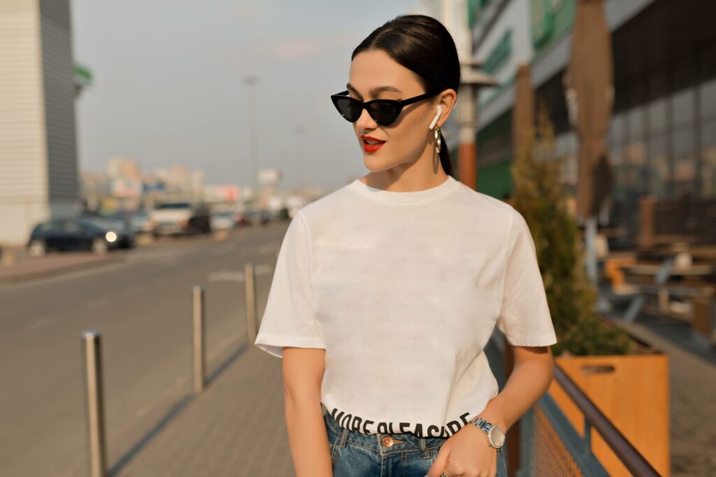 Fashionable girl in white tee and black glasses, enjoying music with red lips against urban backdrop