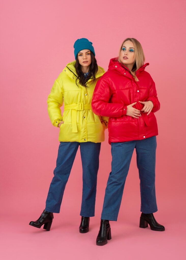 Two stylish young women pose against a pink background, wearing eye-catching winter down jackets in bright red and yellow colors.