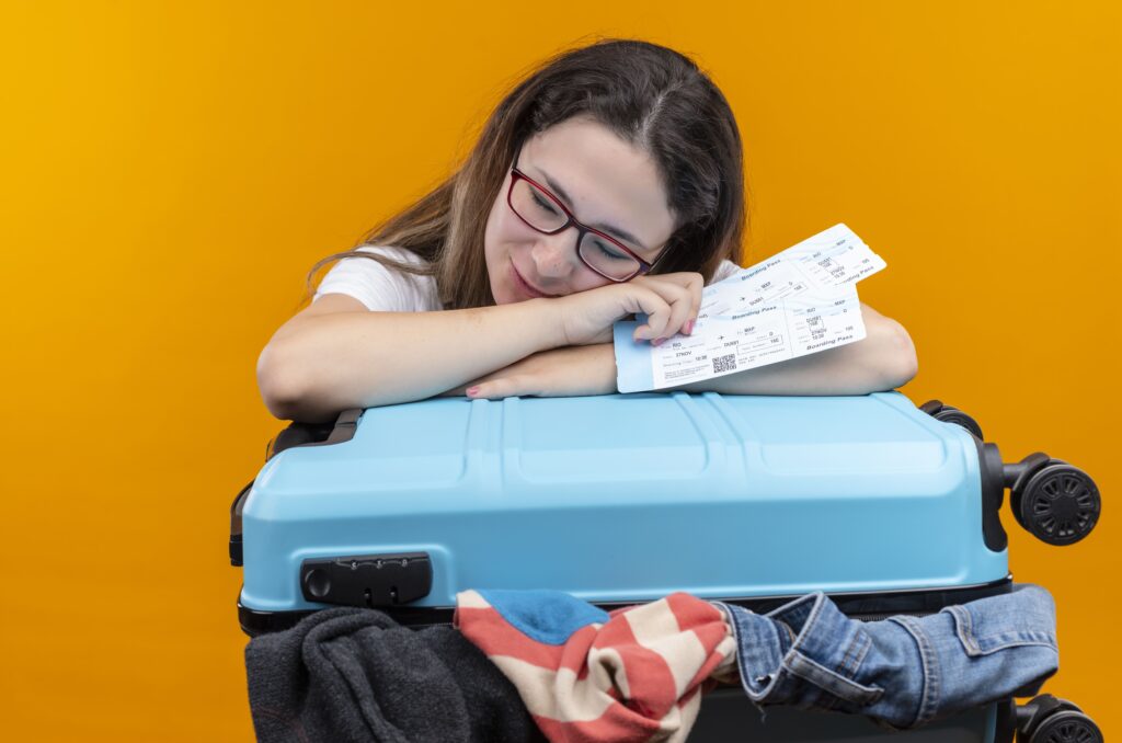 Young traveler woman in white t-shirt standing with suitcase full of clothes holding air tickets leaning head on suitcase sleeping over orange wall