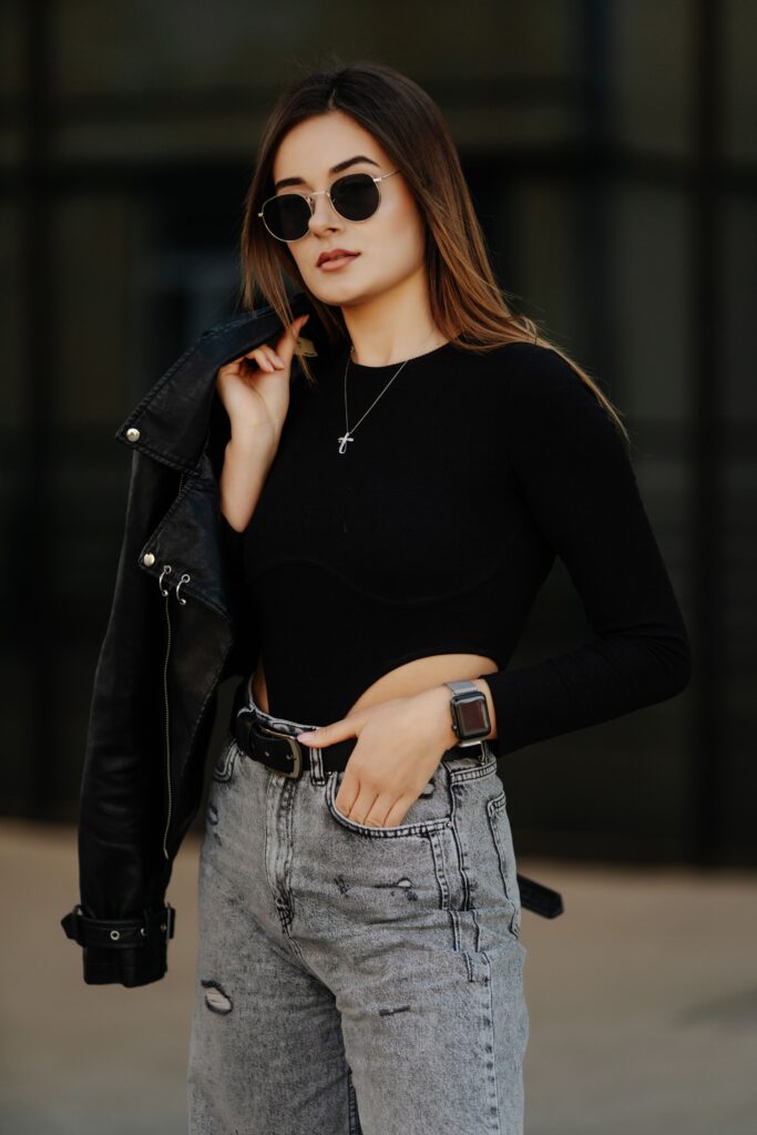 Young woman in sunglasses, hat, and black leather jacket posing outdoors, developing her fashion sense