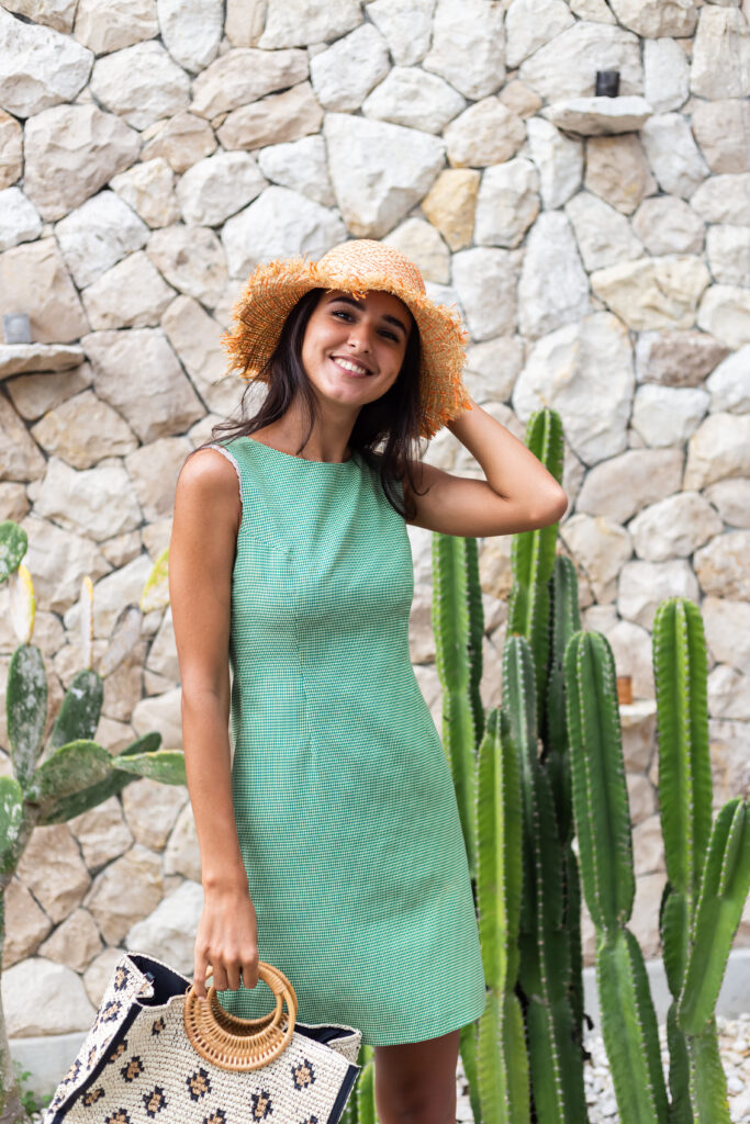 Portrait of a joyful, stylish woman in an elegant green summer dress and straw hat, holding a bag, posing near a white stone wall and cactus.