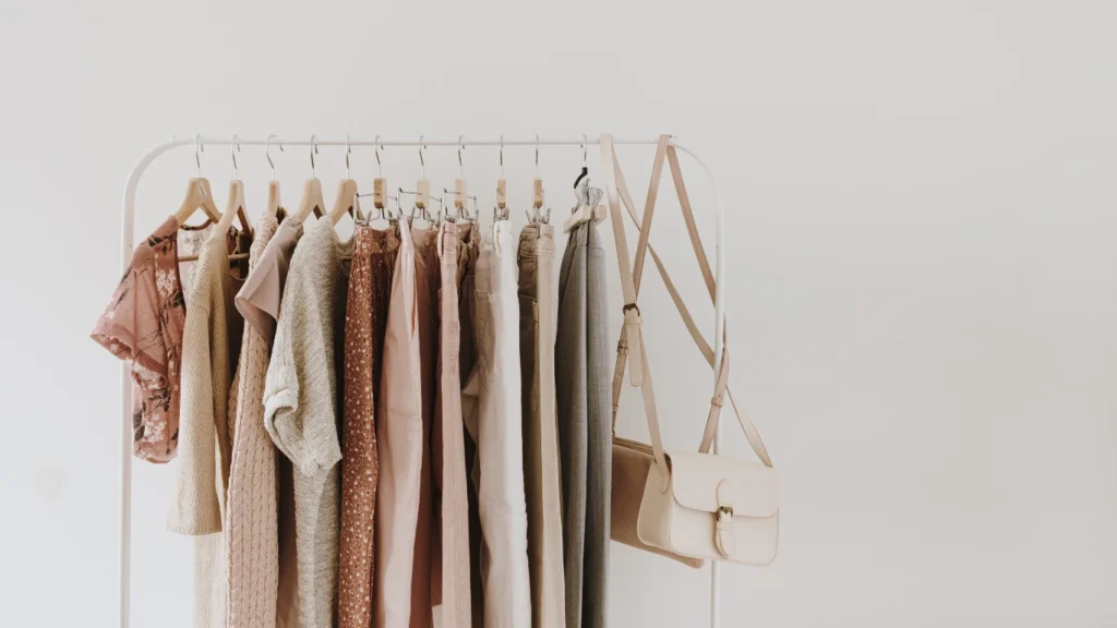 Collection of women's minimalist fashion items in pastel colors, including blouses, sweaters, pants, jeans, t-shirts, and handbags, displayed on a hanger against a white background.
