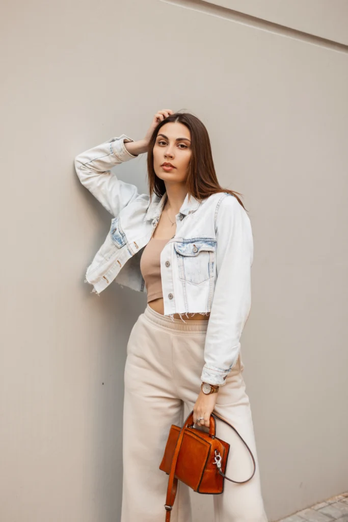 Trendy young beautiful woman with fashion blue denim jacket top pants and handbag stands and poses near a gray wall