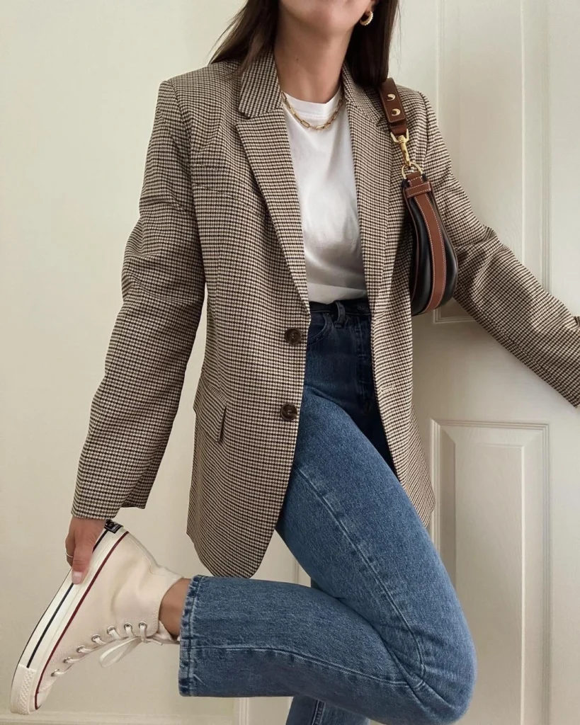 Image of a woman exhibiting effortless chic style, wearing medium-wash denim jeans, a plaid blazer, cream Converse shoes, a white t-shirt, and accessorizing with a shoulder bag.
