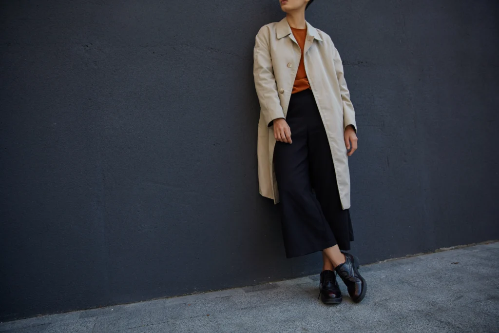 Young woman showcasing Seasonal Minimalist Wardrobe Transition with black culottes, foxy sweater, and beige trench against an urban black wall.