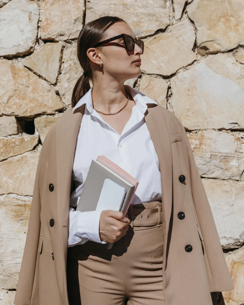 Minimalist woman wearing a layeres white blouse, beige blazer and a beige pants holding books wearing sunglasses