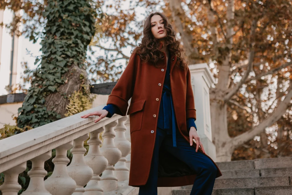 Beautiful stylish smiling skinny woman with curly hair walking in street stairs dressed in warm brown coat and blue suit, autumn trendy fashion street style