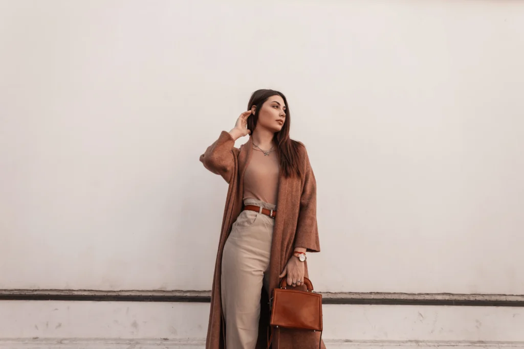 Young woman in elegant coat and pants with brown handbag near a vintage white building.