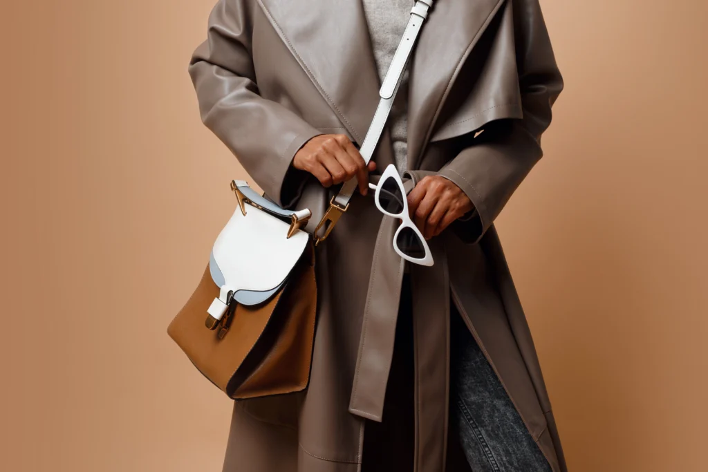 Details. black woman wearing grey leather coat , posing on beige background. brown bag and white sunglasses in hands. autumn or winter fashion concept.