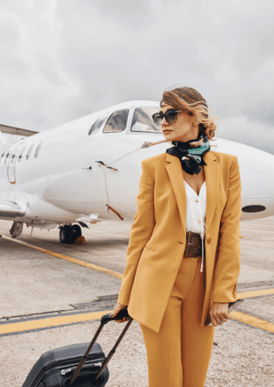 An image of a young woman dressed in stylish yellow airplane outfits stands outdoors near an airplane, holding her luggage