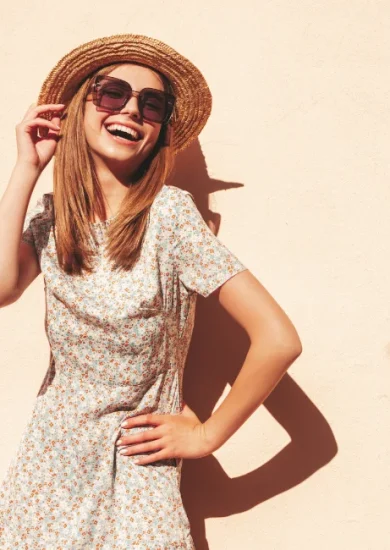 "A woman smiling and wearing a floral dress, wide-brimmed straw hat, and oversized sunglasses, showcasing elements of a summer capsule wardrobe."