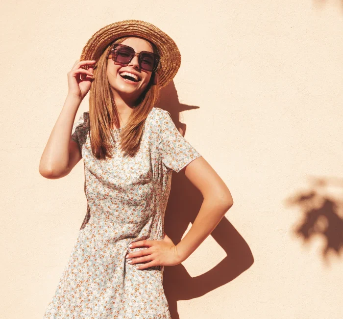 "A woman smiling and wearing a floral dress, wide-brimmed straw hat, and oversized sunglasses, showcasing elements of a summer capsule wardrobe."