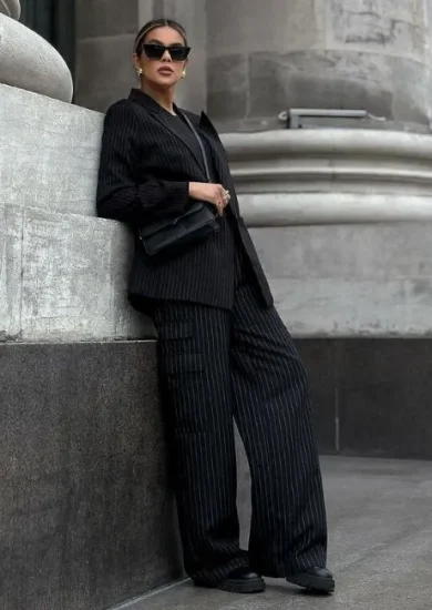 Fashionable woman in a pinstripe suit and black pants outfits leaning against a stone wall.