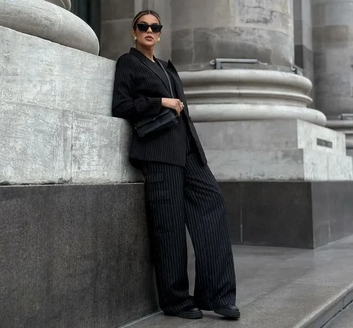 Fashionable woman in a pinstripe suit and black pants outfits leaning against a stone wall.