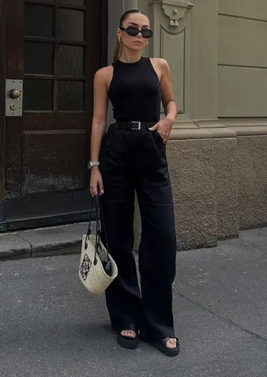 A stylish woman wearing women's black pants, paired with a sleeveless black top, black sandals, and a chic belt, accessorized with sunglasses and a designer handbag, standing confidently in an urban setting.