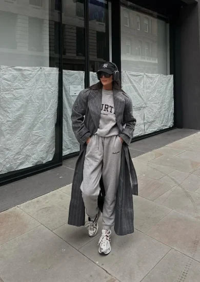 Girl in athleisure fashion, sporting a casual yet chic ensemble with baggy sweats, a gray shirt, a dark gray trench coat, and white sneakers, complemented by sunglasses and a hat.