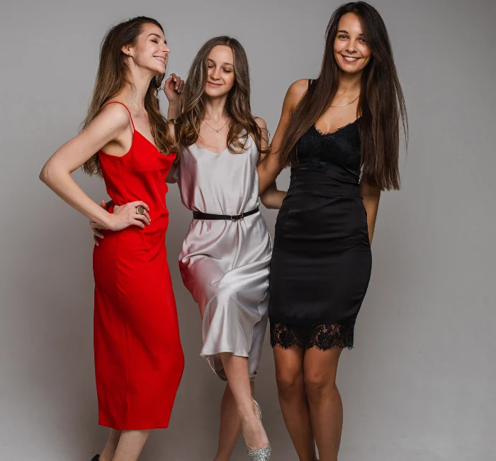 Stylish Types of Dress Codes: Positive Young Girls Friends Posing in Charming Feminine Evening Dresses on Gray Studio Background