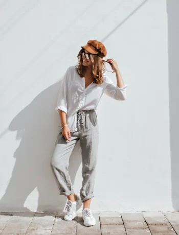 Stylish girl in gray pants and white cotton blouse posing near white wall, showcasing a Seasonal Minimalist Wardrobe Transition look. Woman in cap and glasses.