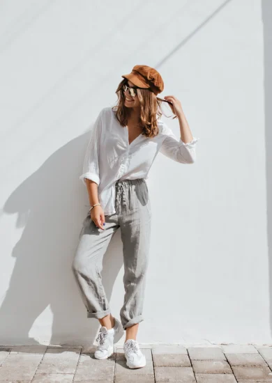 Stylish girl in gray pants and white cotton blouse posing near white wall, showcasing a Seasonal Minimalist Wardrobe Transition look. Woman in cap and glasses.