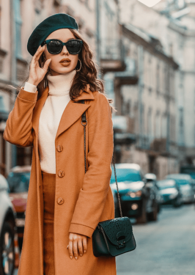 A stylish young woman showcasing her personal style in an outdoor fall portrait, wearing trendy sunglasses, a camel-colored coat, turtleneck, and a textured leather shoulder bag, strolling through the streets of a European city.