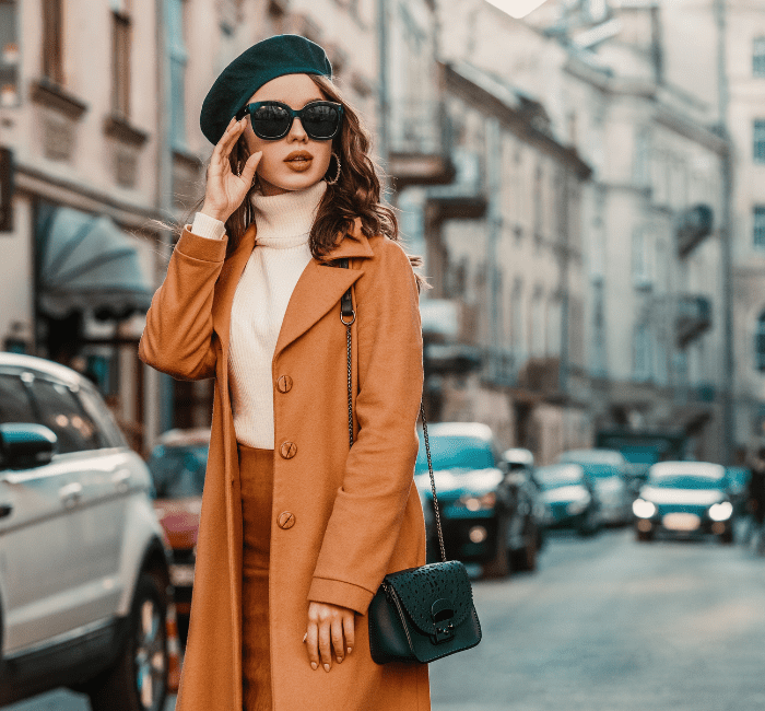 A stylish young woman showcasing her personal style in an outdoor fall portrait, wearing trendy sunglasses, a camel-colored coat, turtleneck, and a textured leather shoulder bag, strolling through the streets of a European city.