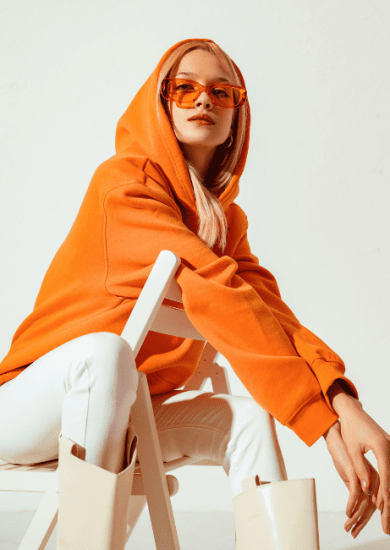 Stylish young blonde woman in a vibrant orange hoodie and colorful sunglasses, striking a pose against a white backdrop in a studio fashion portrait