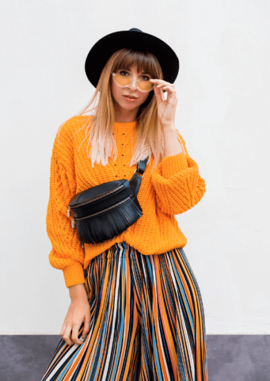 Cheerful brunette woman defying fashion rules, standing on white background in trendy orange sweater and multicolor striped culottes