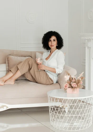 Woman with curly hair sits on a sofa with bare feet and holds a glass of water enjoying minimalist living