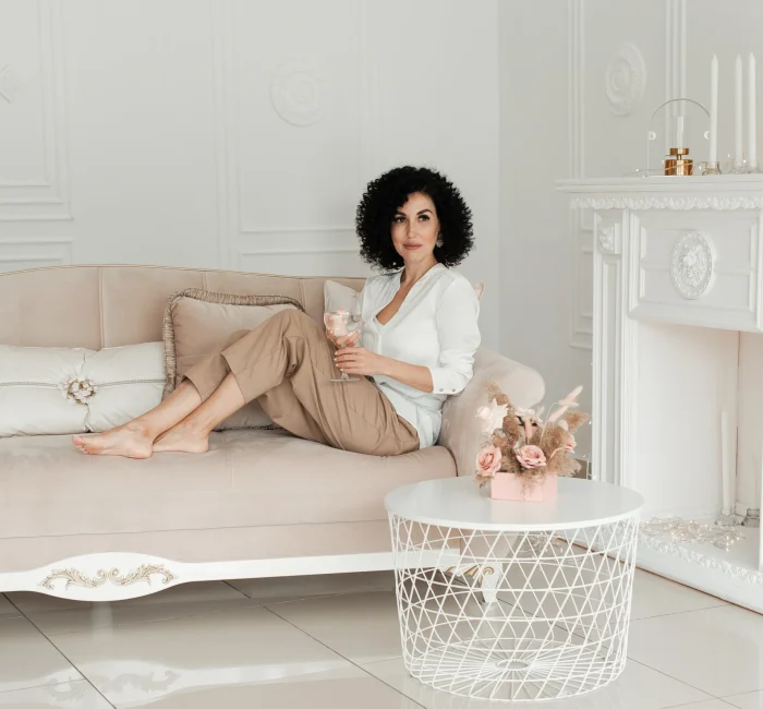 Woman with curly hair sits on a sofa with bare feet and holds a glass of water enjoying minimalist living